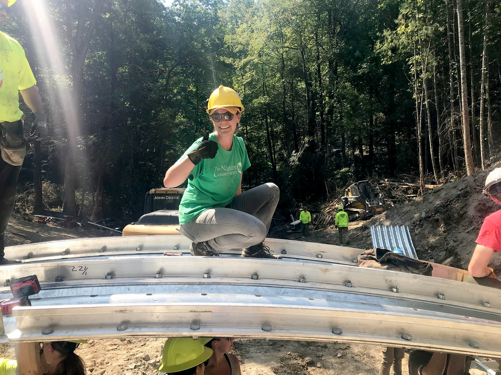 Smiling young woman in green t-shirt and hard hat stands on top of construction equipment