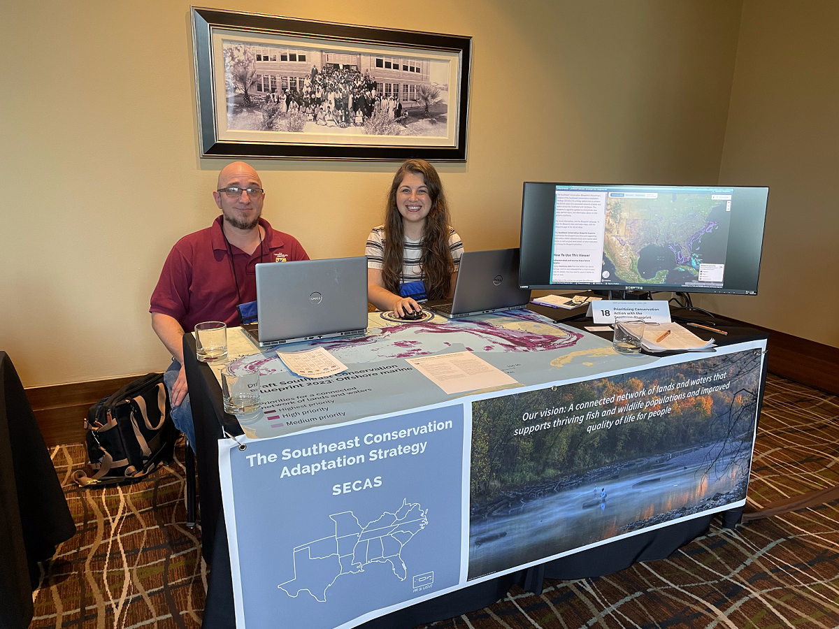 Smiling man and woman sit behind table with banner and large screen showing Blueprint map.