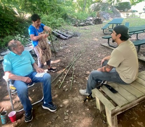 Young man sits on a bench watching an older woman demonstrate rivercane techniques.