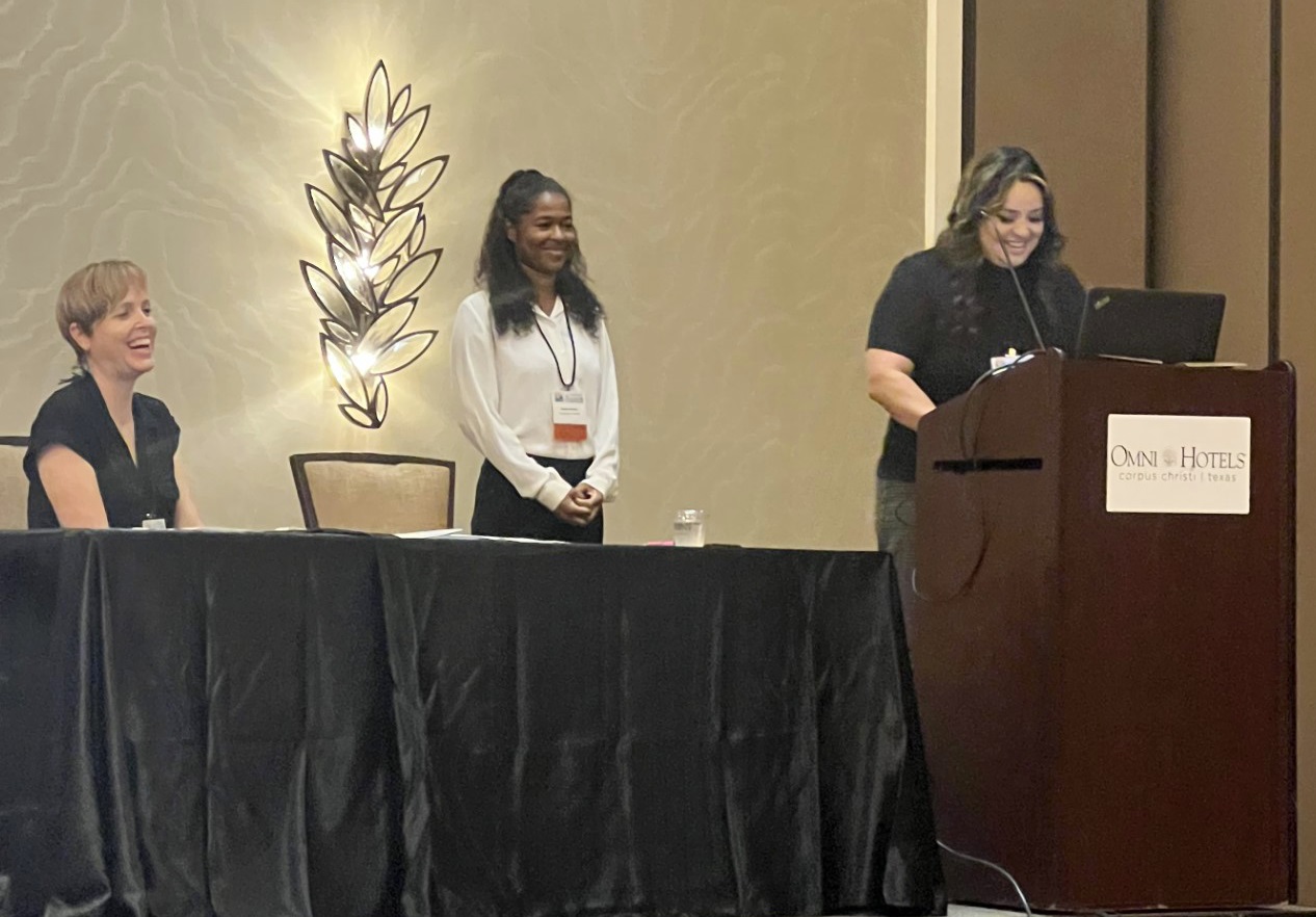 Photo of Tanya standing at the podium with Jaiere and Amanda on the platform, smiling and laughing