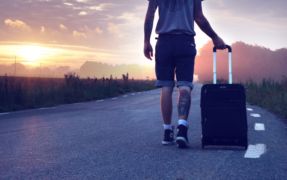 Man walks along a scenic road toward the sunset, pulling a black rolling suitcase.