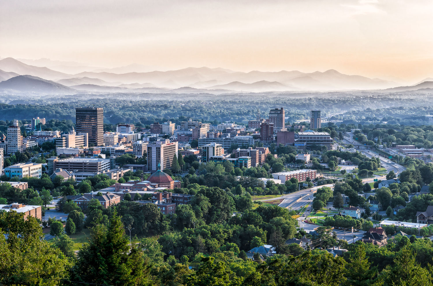 Photo of the Asheville city skyline with tall buildings among green trees with mountains in the background.