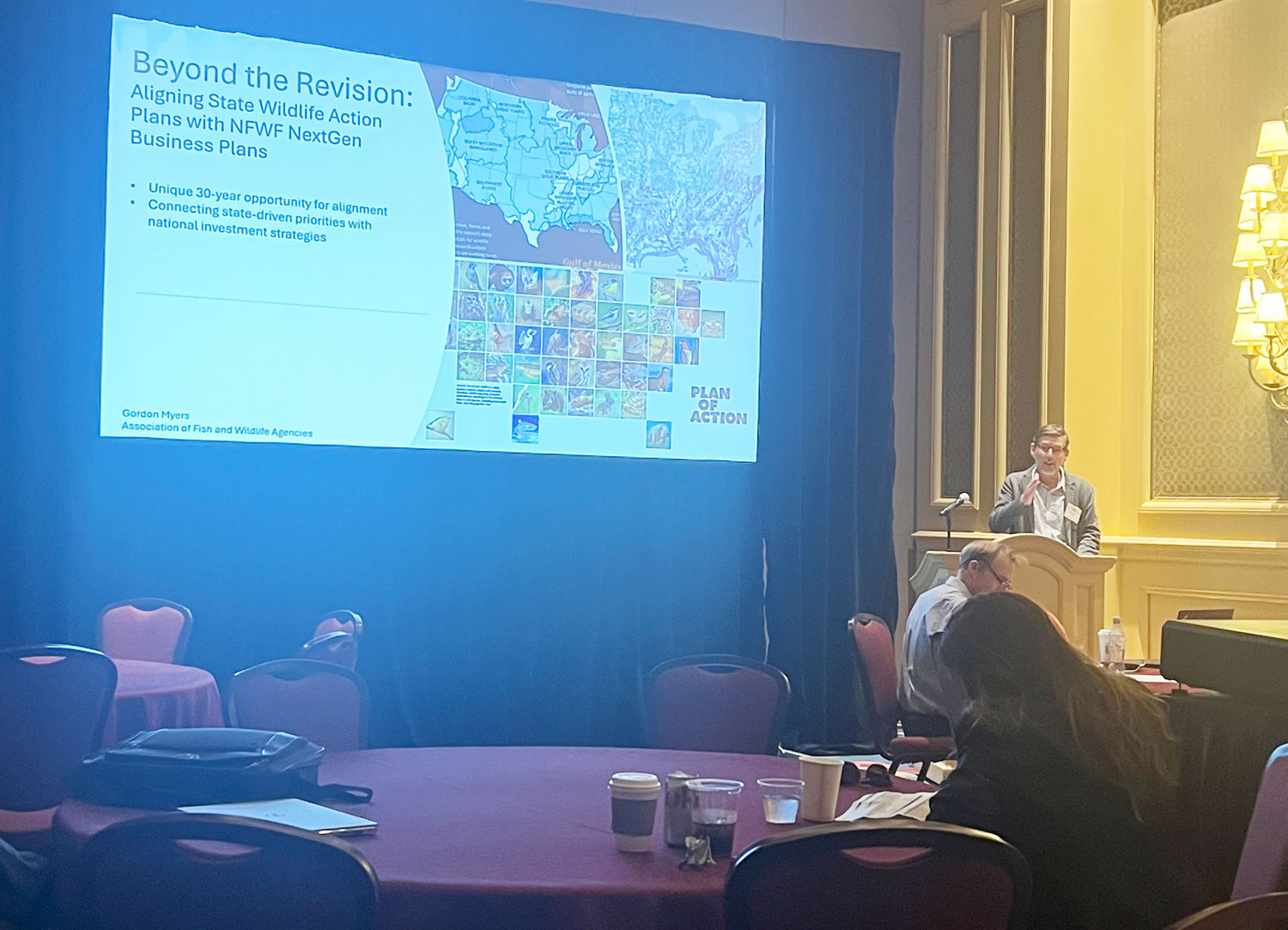 Photo of a man standing at a podium in front of a powerpoint slide on a projector screen.