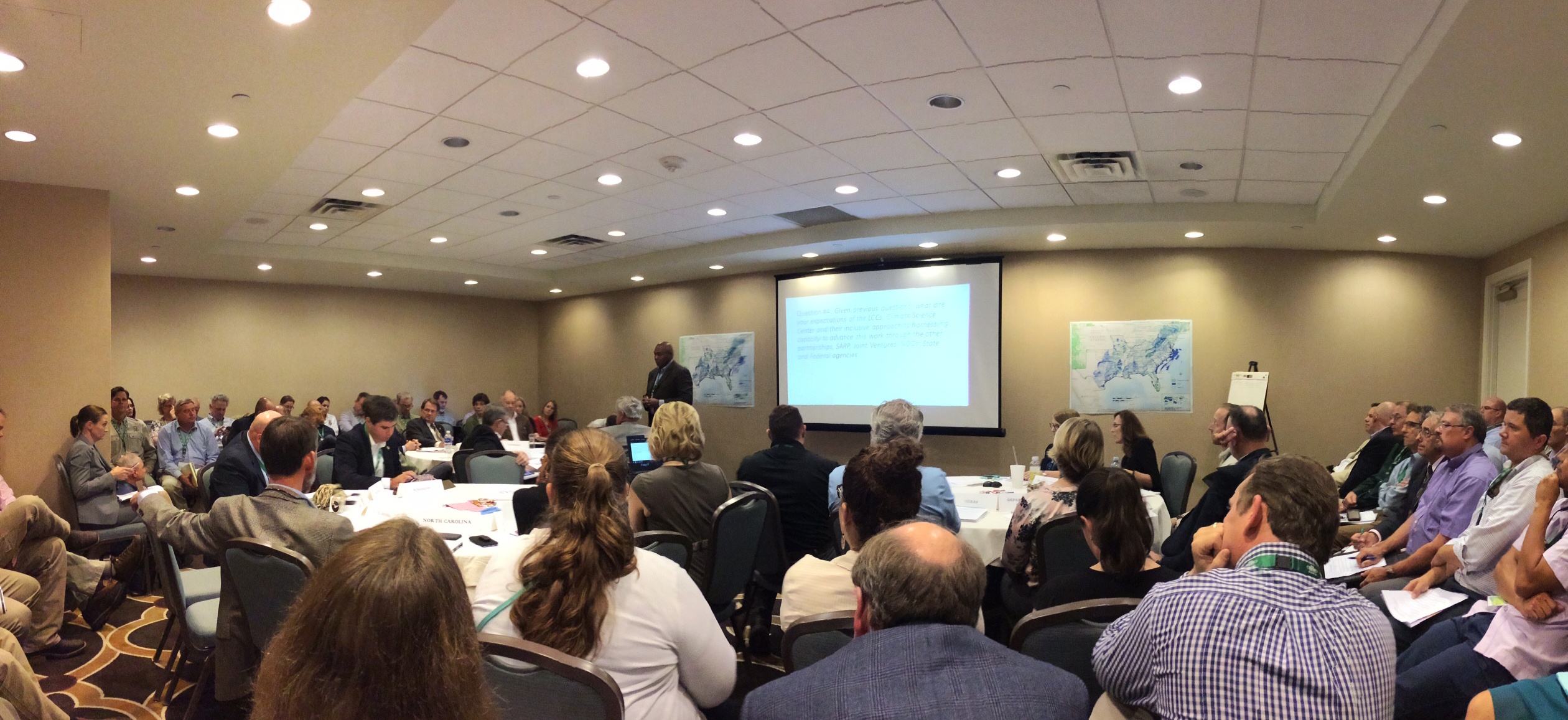 Panoramic image of the SECAS leadership summit, showing a packed conference room with a projector screen in the front, along with several maps of the Southeast Blueprint on the walls and an easel for notes.