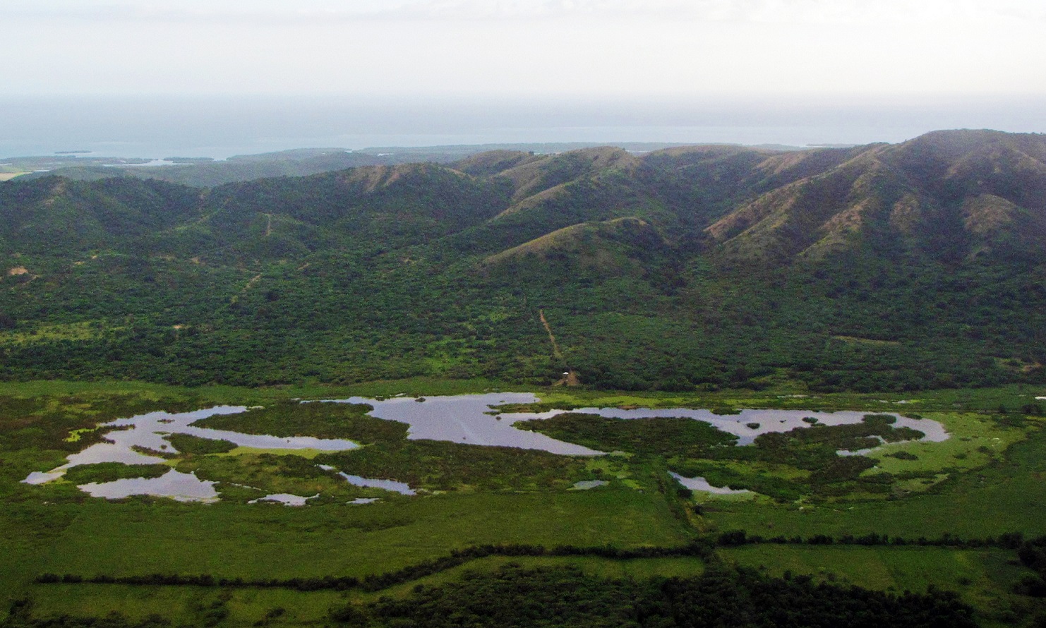 View of a green scenic landscape with forested mountains in the background and a lagoon in the foreground.