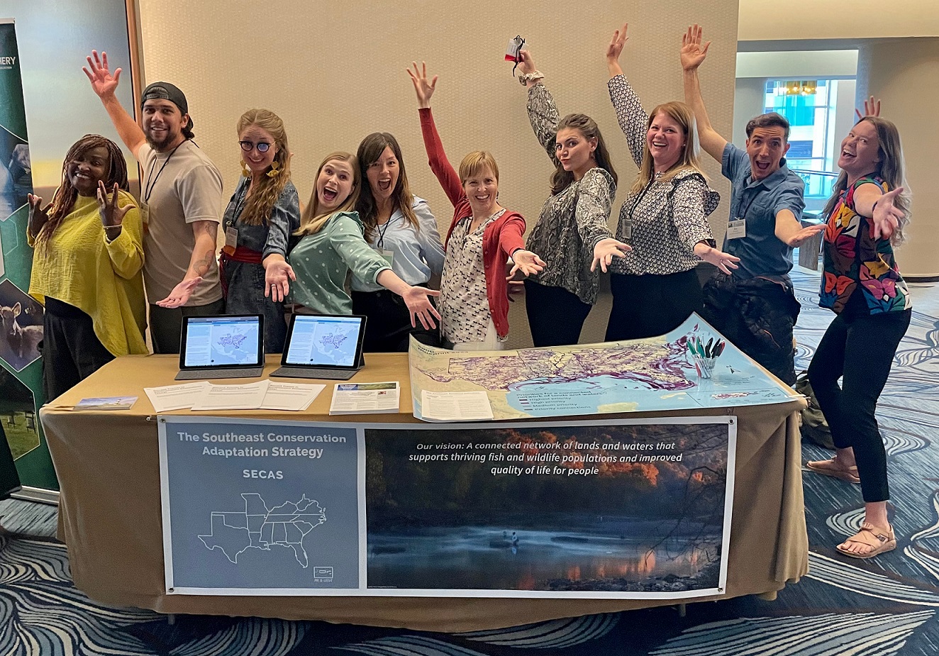 Silly group photo of SECAS staff at SEAFWA standing behind the SECAS booth. A table with a SECAS banner and iPads showing maps of the Blueprint with 10 smiling people posing behind it.