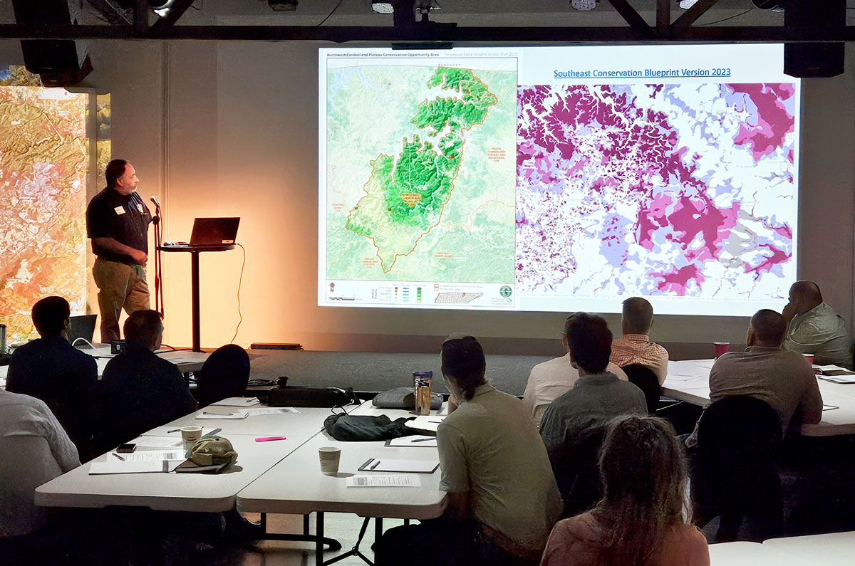 Man stands at podium presenting next to a screen showing maps of Tennessee, one of which is the purple priorities of the Southeast Conservation Blueprint.